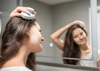 Young woman doing self hair scalp massage with scalp massager or hair brush for hair growth stimulating at home bathroom. Reflected view of the mirror