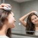 Young woman doing self hair scalp massage with scalp massager or hair brush for hair growth stimulating at home bathroom. Reflected view of the mirror