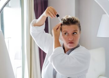 Middle-aged woman applies hair serum with a dropper to her scalp in front of a mirror