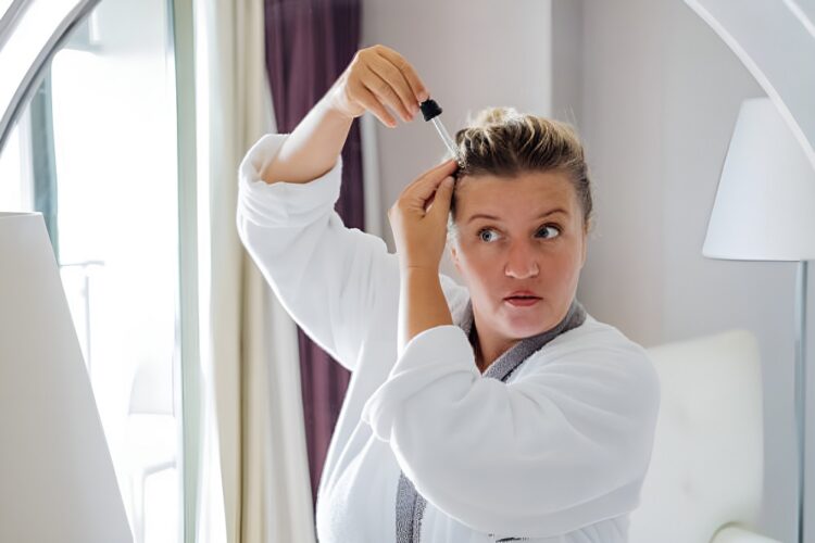 Middle-aged woman applies hair serum with a dropper to her scalp in front of a mirror